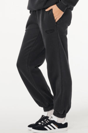 A person wearing Rip Curl Varsity Sports Track Pant, a black sweatshirt, white socks, and black Adidas sneakers with white stripes stands with one hand in a pocket against a white background.