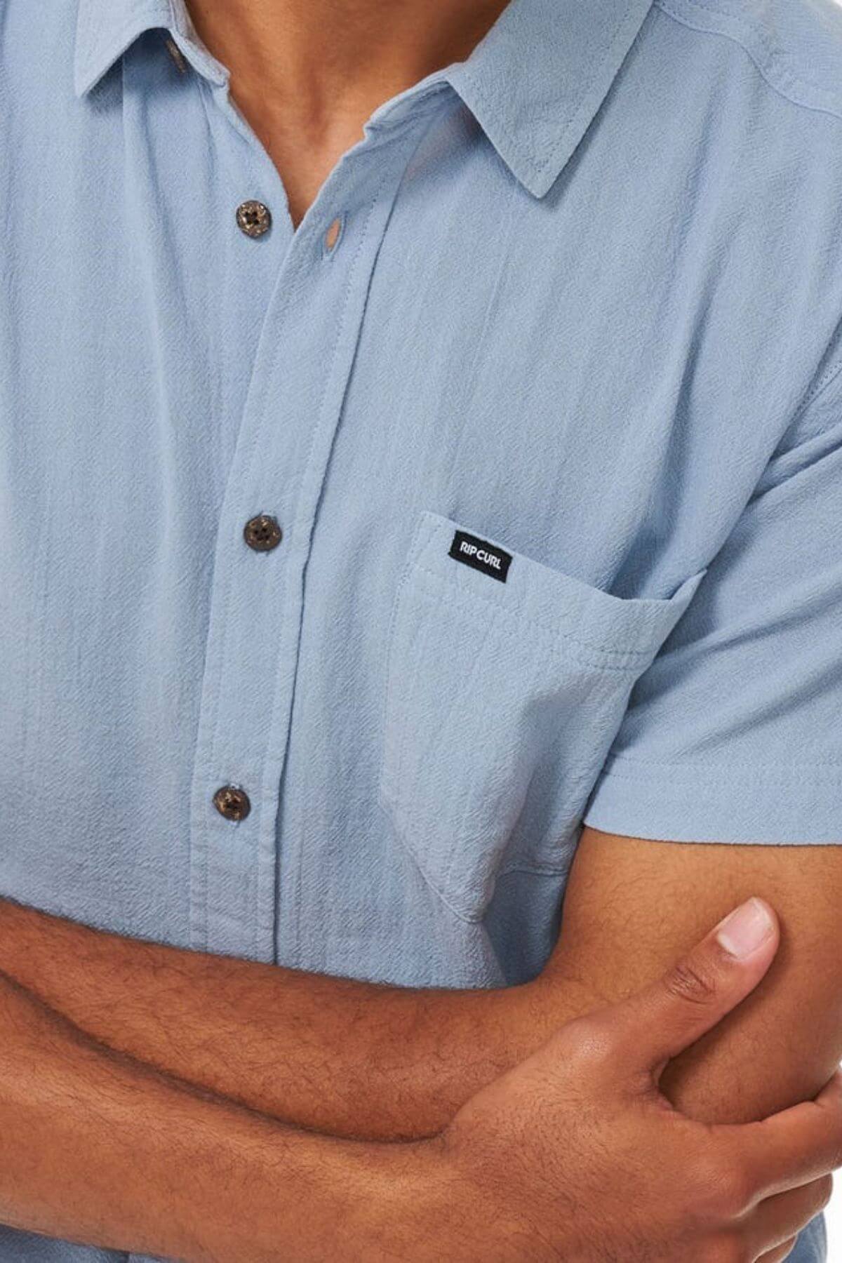 A person with crossed arms wears the Rip Curl Washed Short Sleeve Shirt in light blue, featuring a front pocket with a small "RIP CURL" tag. Only the torso and part of their arms are shown.