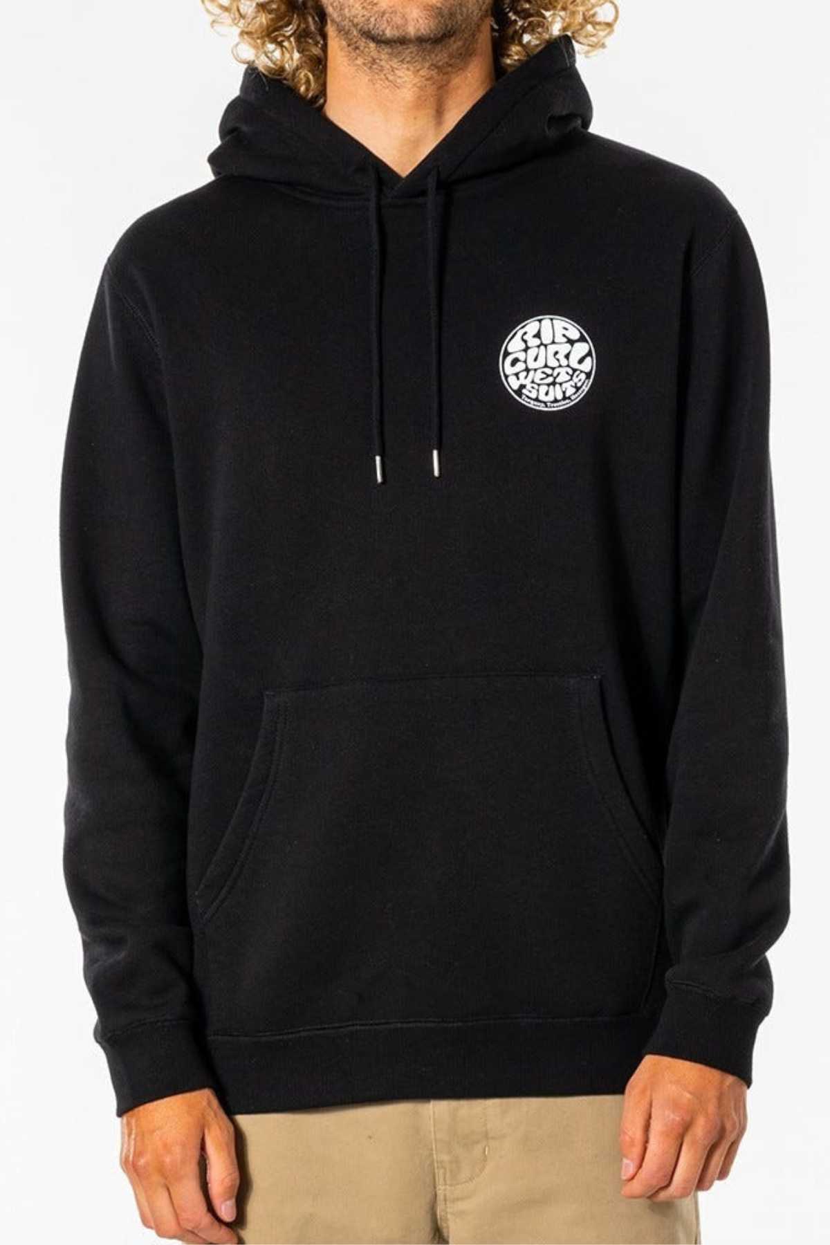 Someone wearing the Rip Curl Wetsuit Icon Hood, a black organic cotton poly fleece hoodie with a white circular chest graphic and kanga pocket, paired with tan pants against a plain light background.