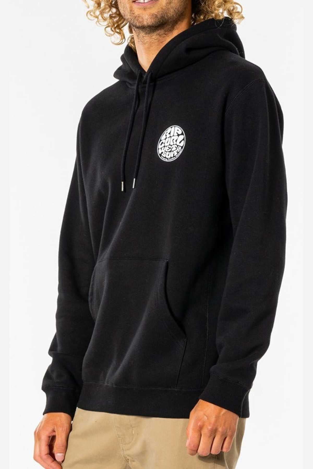 Someone wearing the Rip Curl Wetsuit Icon Hood—a black hoodie with kanga pocket and small white circular chest logo—pairs it with tan pants. Their curly hair is visible, but their face is partially out of frame.