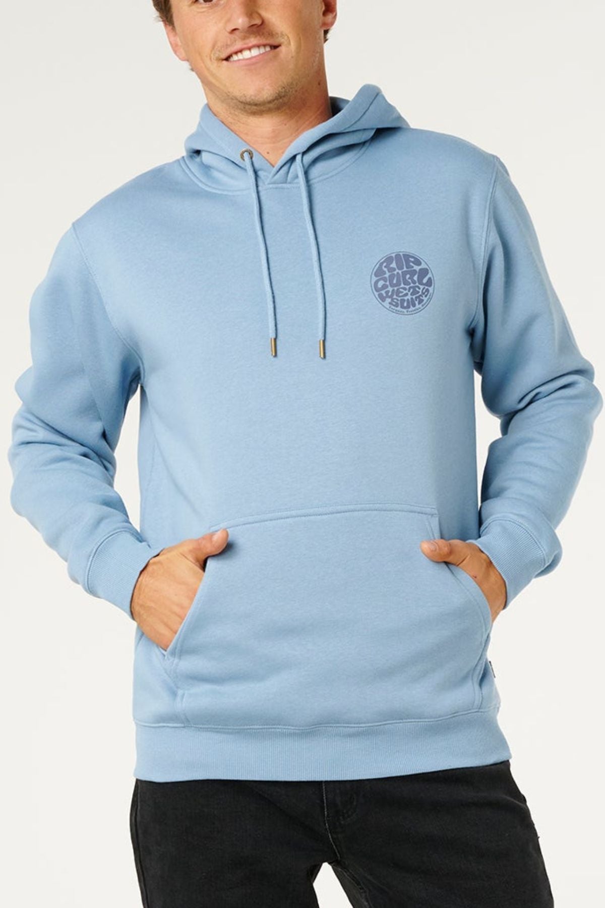 A smiling person wears the Rip Curl Wetsuit Icon Hood in light blue, featuring the signature Wettie Logo on the left chest with both hands in the front pocket, standing against a plain light background.