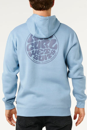 A person faces away from the camera, wearing a casual light blue Rip Curl Wetsuit Icon Hood featuring the iconic Wettie Logo and "Rip Curl Wetsuits" with locations printed on the back.