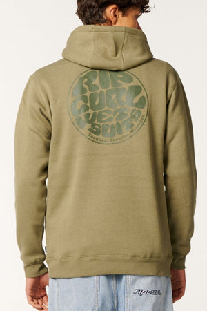 A person in light blue jeans stands with their back to the camera, wearing an olive green Rip Curl Wetsuit Icon Hood made from organic cotton fleece with a circular logo on the back and a cozy hood.