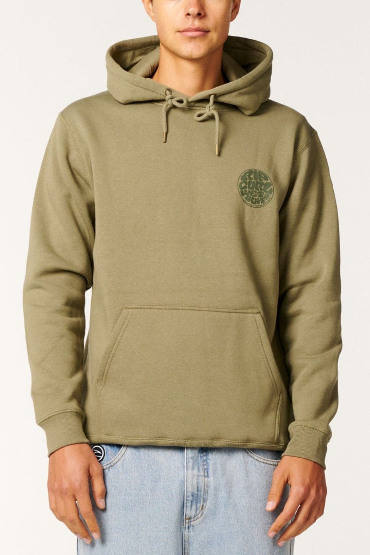 A person wearing the Rip Curl Wetsuit Icon Hood in light olive green organic cotton fleece, with a small round green chest graphic, paired with light blue jeans, stands against a plain background.