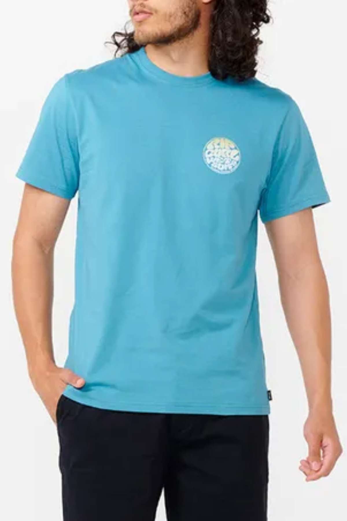 A person with long, curly hair is wearing a Rip Curl Wetsuit Icon Tee in blue, featuring a small ocean print graphic on the chest, paired with black pants. The circular multicolored design adds flair as they pose with their left hand in their pocket, looking off to the side.