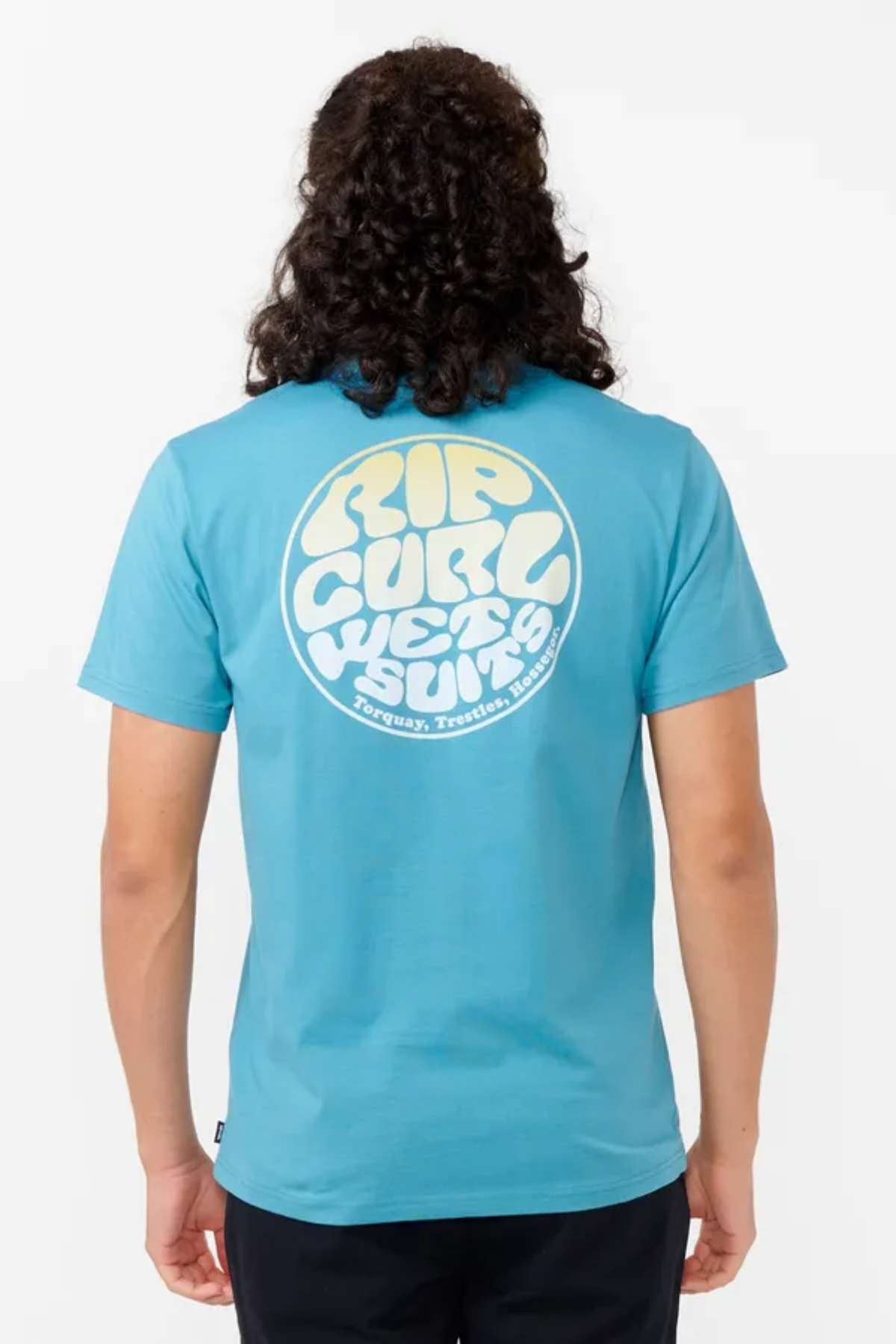 A person with long, curly hair is wearing the Rip Curl Wetsuit Icon Tee made from organic cotton. The shirt is a blue t-shirt featuring a retro-style ocean print logo on the back, elegantly framed at the bottom by the words "Torquay, Trestran, Hossegor.