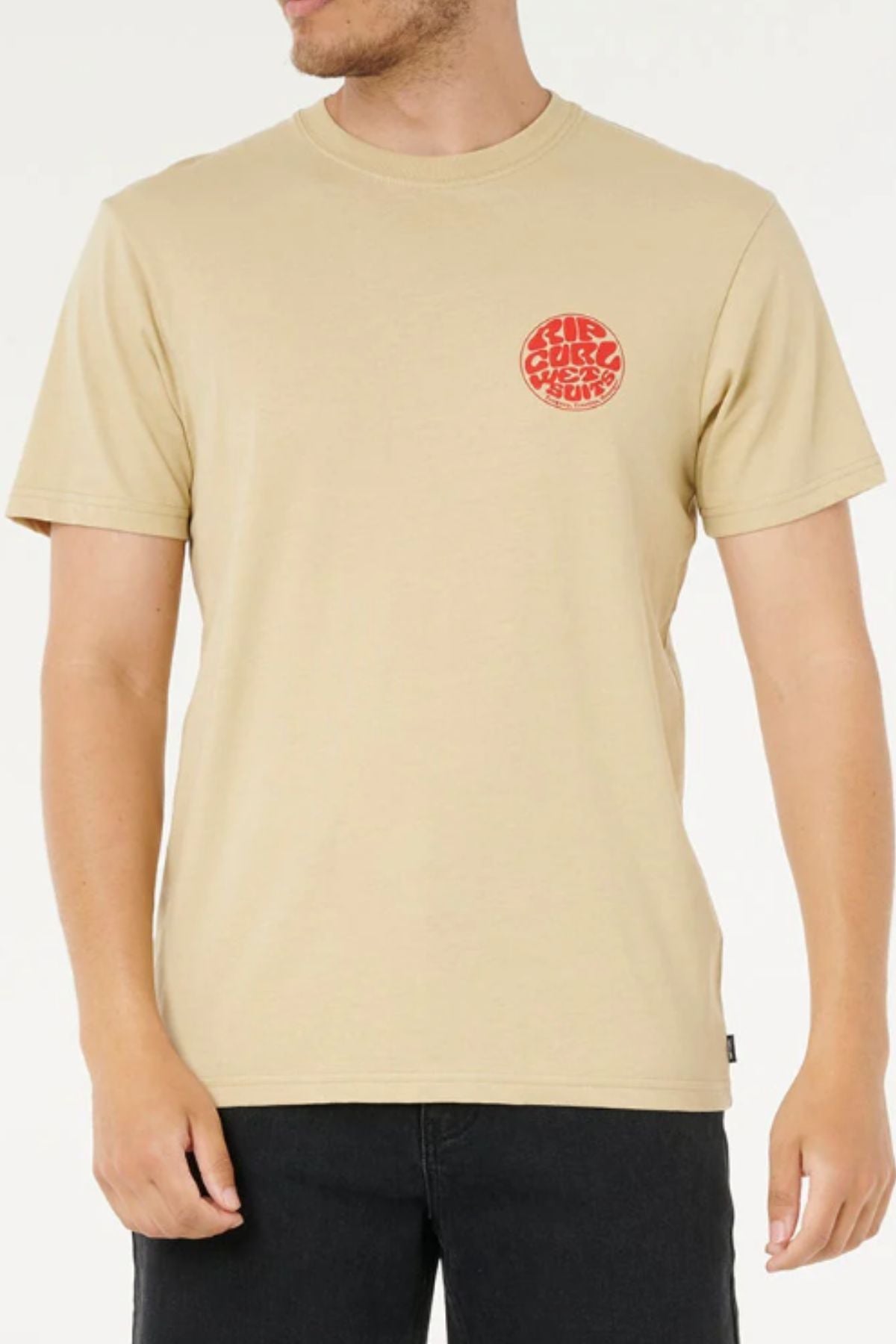 A person wearing the Rip Curl Wettie Icon Tee, a beige 100% cotton T-shirt with a small red circular graphic on the left chest, paired with black jeans and standing against a plain light background.