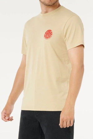A man wearing the Rip Curl Wettie Icon Tee, a beige 100% cotton T-shirt with a small red circular graphic on the left chest, and black pants, stands against a plain white background.