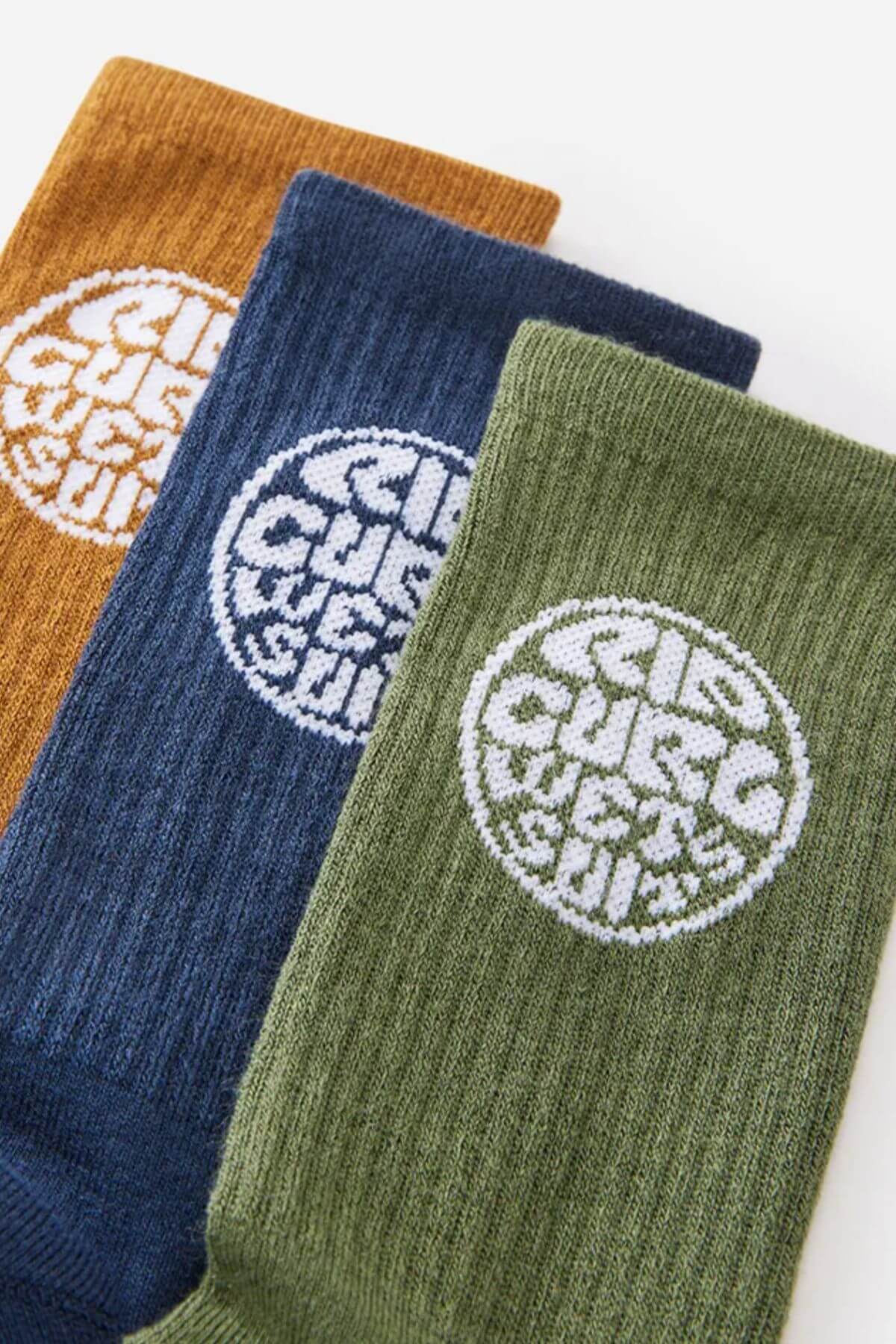 The Rip Curl Wetty Crew Sock 3 Pack features mustard, navy, and olive ribbed socks with a white circular logo. The logo text is unreadable, and each sock provides comfortable arch support for all-day wear.