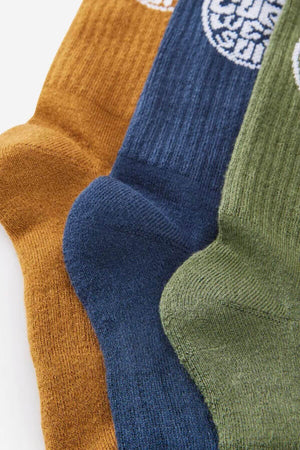 Close-up of three overlapping Rip Curl Wetty Crew socks in ochre, navy blue, and olive green on a white background. Each ribbed sock has a white circular logo near the top and provides arch support.