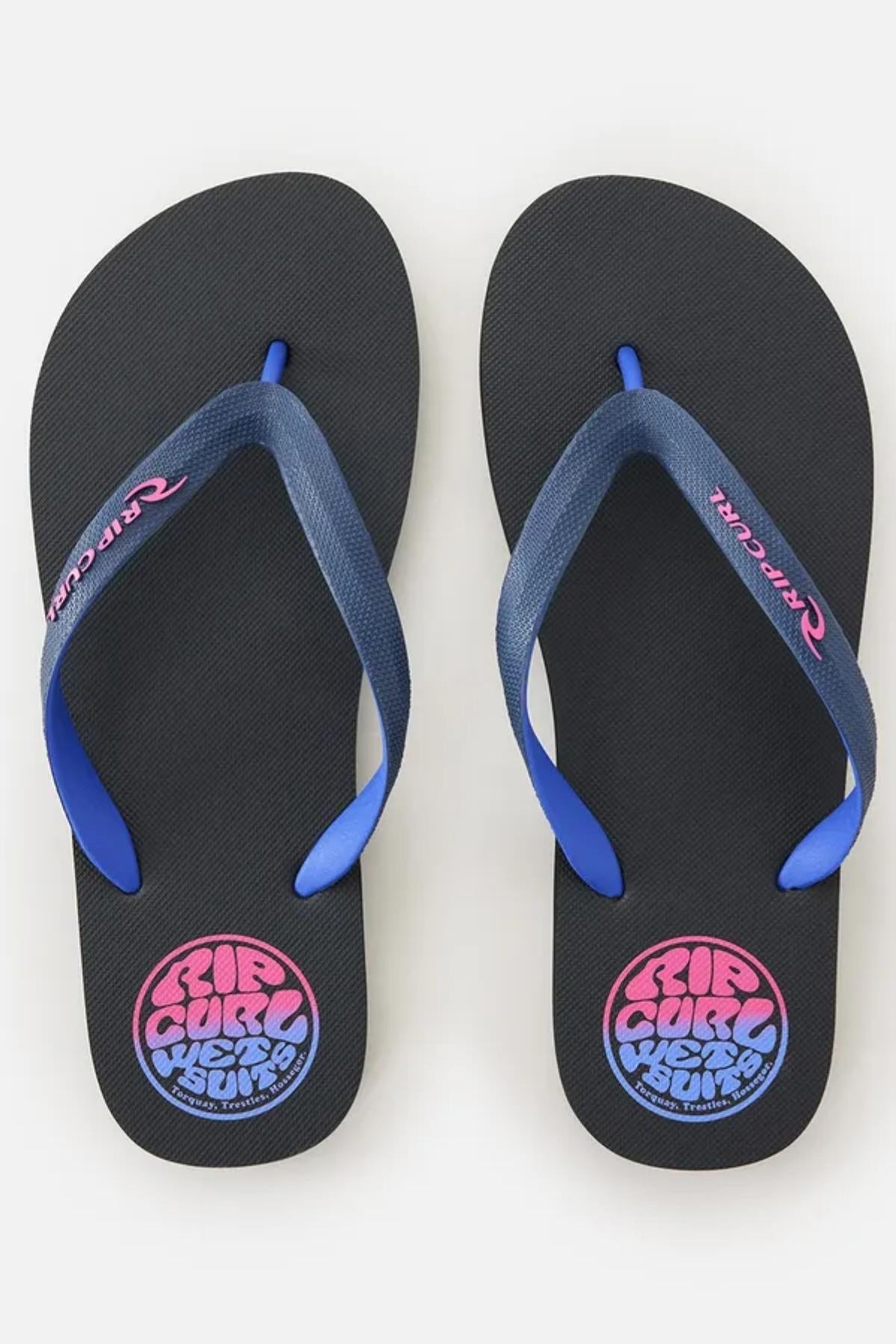 The Rip Curl Wetty Fade Bloom Thongs are black flip-flops with blue straps, adorned with the "Rip Curl" logo in pink and blue on the straps and a circular "Rip Curl" logo near the heel area of each sandal. The background is plain white.