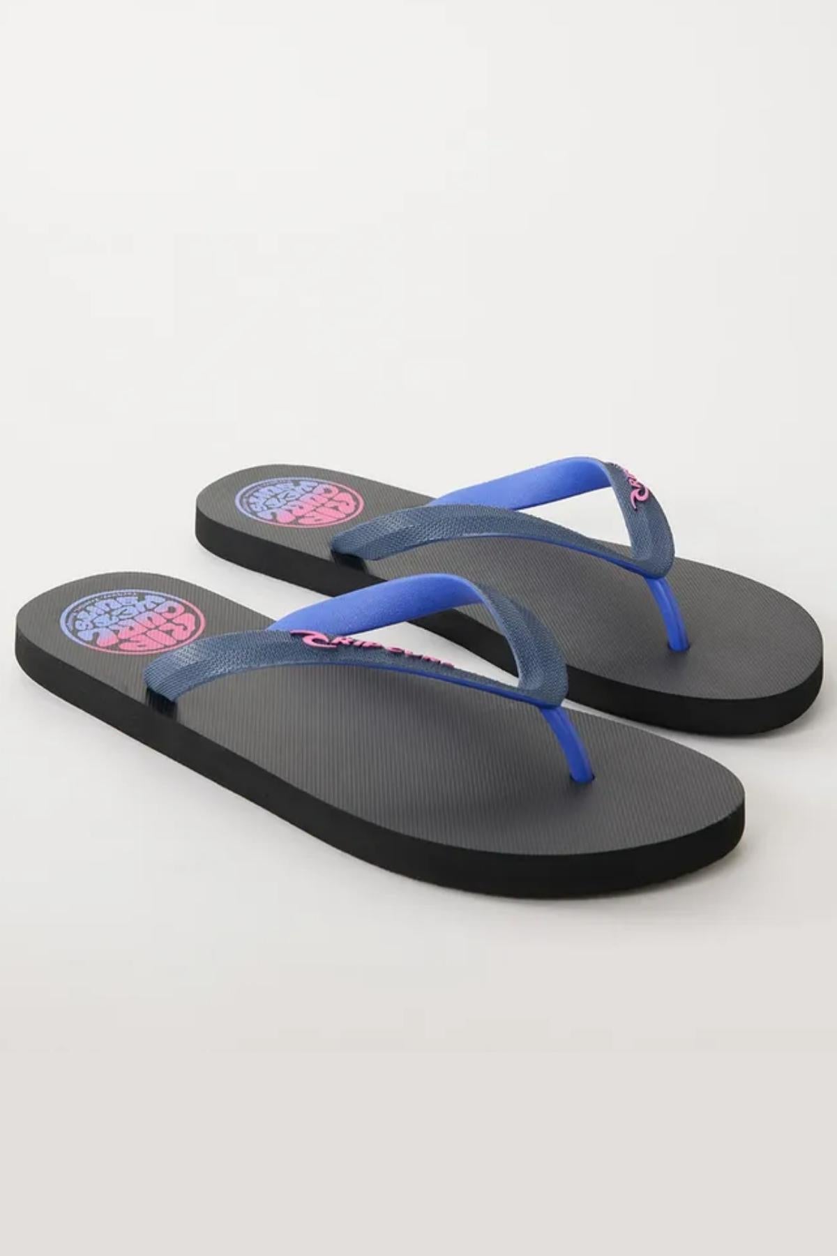 The Rip Curl Wetty Fade Bloom Thongs are black flip-flops featuring dark blue straps. The left strap is emblazoned with "Quiksilver" in pink. A circular pink and blue logo design adorns the insole near the heel area. With flat soles, these flip-flops boast a minimalistic and casual aesthetic.