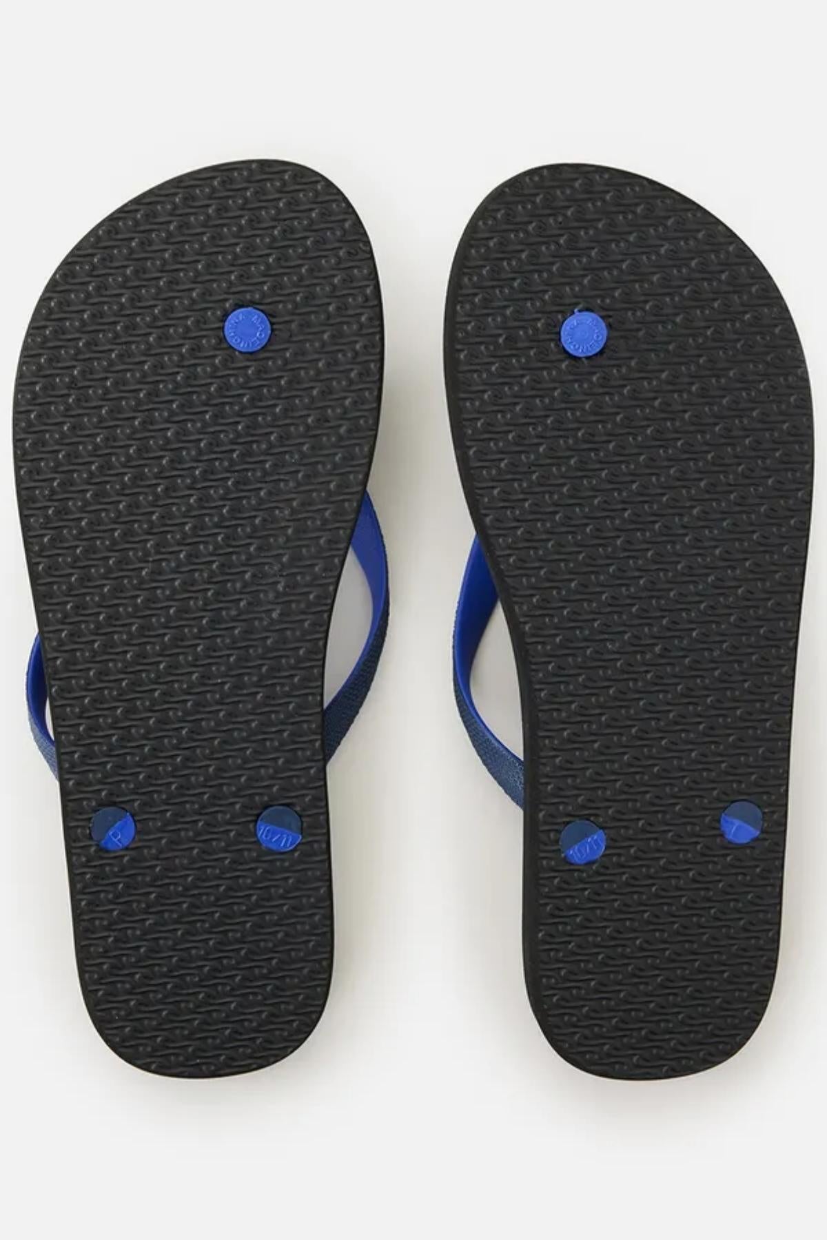 The image shows the bottom view of a pair of Rip Curl Wetty Fade Bloom Thongs, featuring black soles and blue straps. The soles have a textured pattern with three blue circular plugs visible on each thong, likely securing the straps to the sole. The background is plain white.