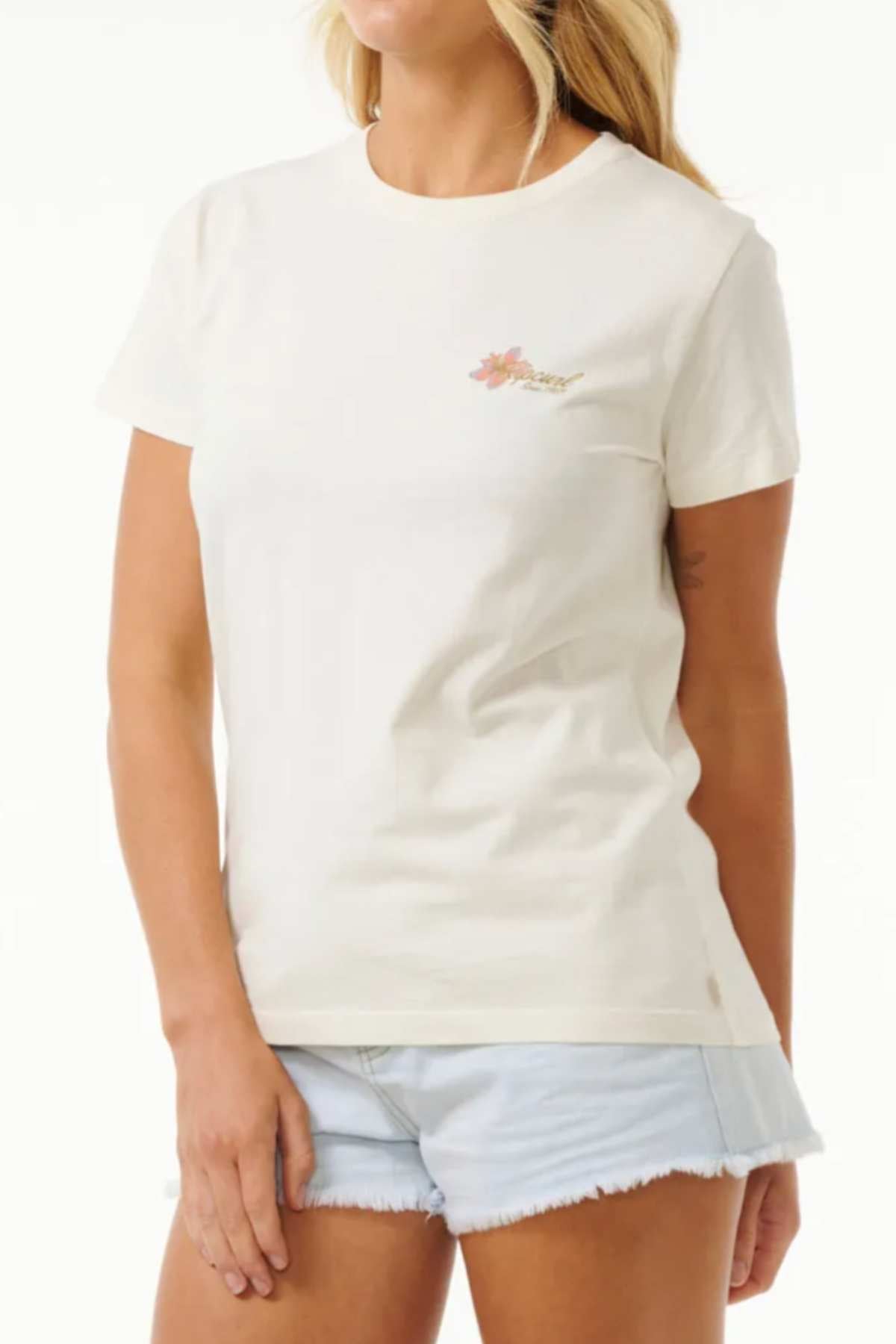 A person with long, blonde hair is standing against a plain background, showcasing a casual wardrobe choice. They are wearing a Rip Curl Wild Bloom Standard Tee, which is short-sleeved and light-colored with a small floral design on the left chest area. The individual pairs this 100% cotton T-shirt with light denim shorts.