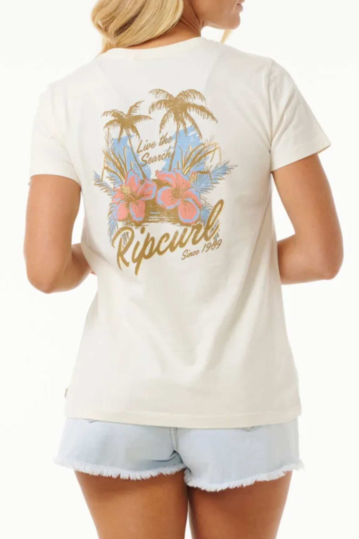 A person with long, blonde hair is seen from behind wearing the 100% cotton Rip Curl Wild Bloom Standard Tee, which showcases a tropical design featuring palm trees, pink hibiscus flowers, and the text "Live the Search Ripcurl Since 1969." The tee pairs perfectly with light denim shorts for a casual wardrobe look.