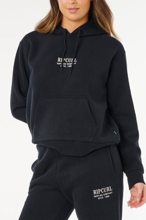 A woman in a black Rip Curl Surf Staple Relaxed Hood with white logos and matching sweatpants stands against a plain white background, showcasing effortless surf-inspired style.