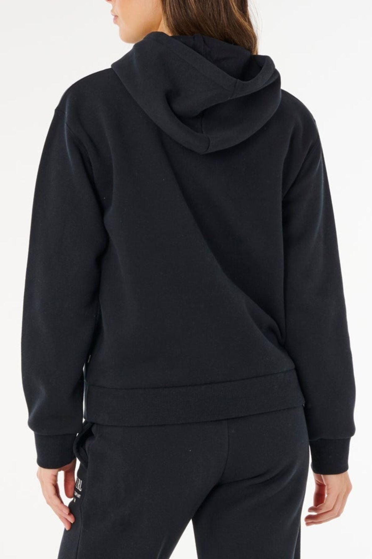 A person is seen from the back, wearing a black Rip Curl Surf Staple Relaxed Hood and black pants, with their head slightly turned left. The hood rests on their upper back, set against a clean white background.
