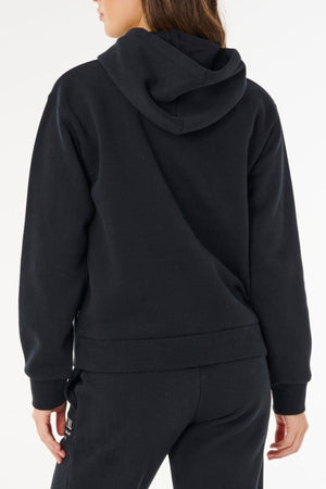 A person is seen from the back, wearing a black Rip Curl Surf Staple Relaxed Hood and black pants, with their head slightly turned left. The hood rests on their upper back, set against a clean white background.