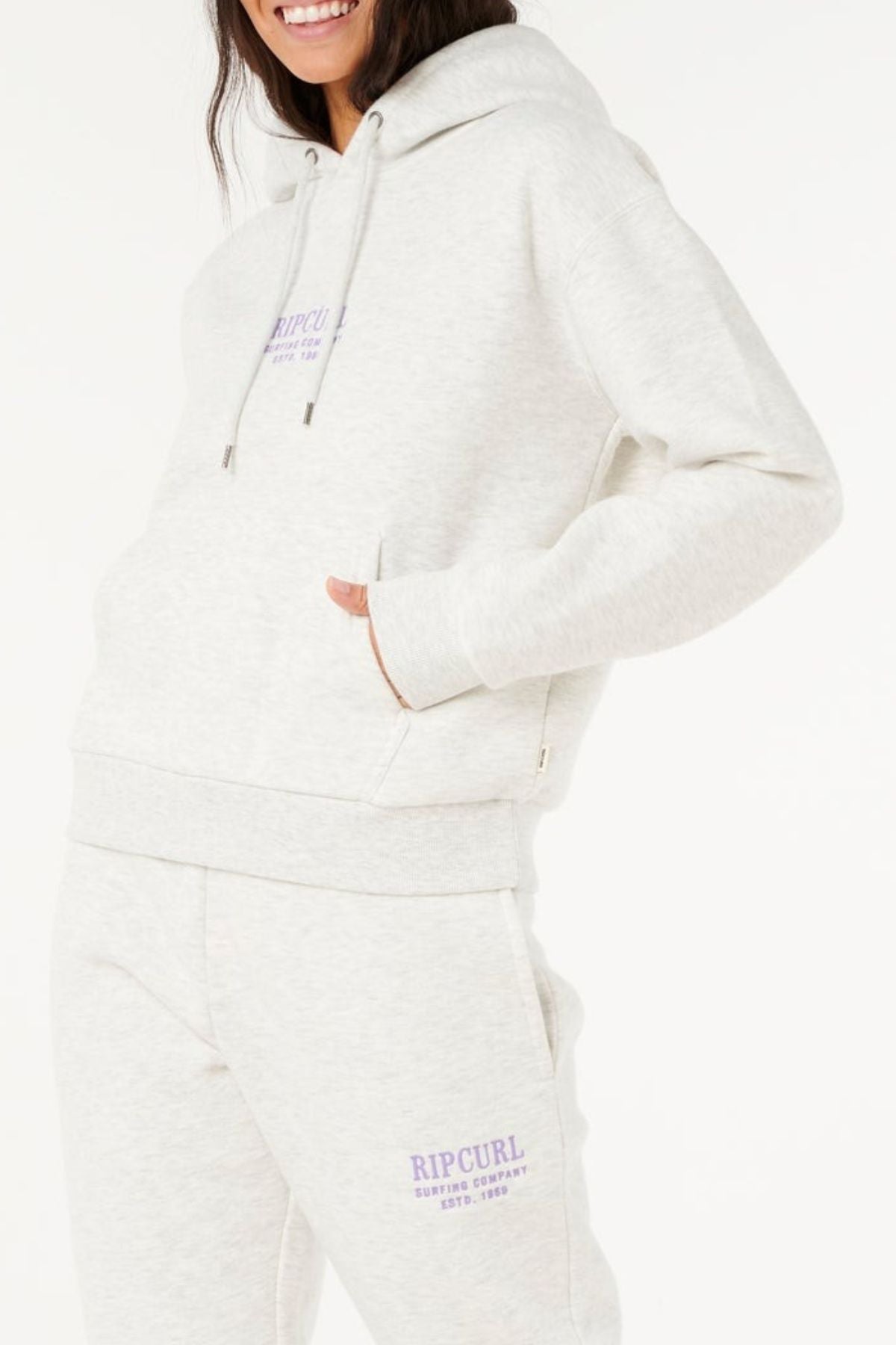 A woman wearing a light gray Rip Curl Surf Staple Relaxed Hood and matching sweatpants with purple logos on the chest and leg, hands in pockets, smiles, showcasing classic surf heritage style.