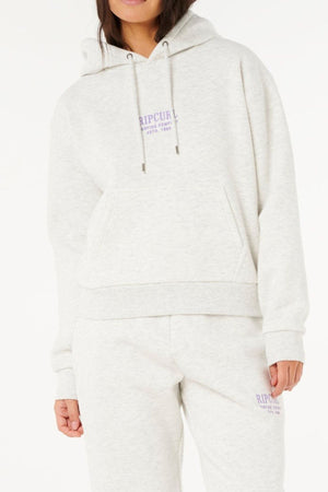A woman wears a light gray Rip Curl Surf Staple Relaxed Hood and matching sweatpants with small purple logos, standing against a white background and showcasing effortless surf-inspired style.
