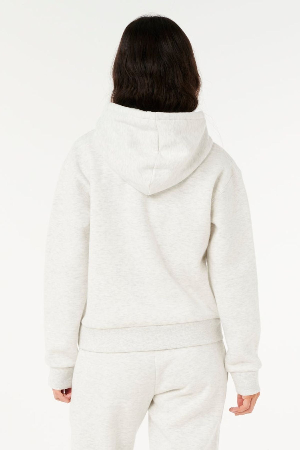 A person with long dark hair, seen from behind, wears a light gray Rip Curl Surf Staple Relaxed Hood and matching sweatpants, reflecting surf heritage against a plain white background.