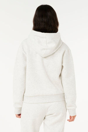 A person with long dark hair, seen from behind, wears a light gray Rip Curl Surf Staple Relaxed Hood and matching sweatpants, reflecting surf heritage against a plain white background.