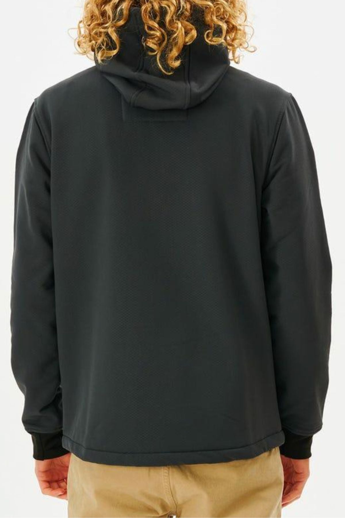 A person with curly blonde hair is shown from the back, wearing the Rip Curl Anti Series Soft Tech Fleece in black and beige pants against a plain light background.