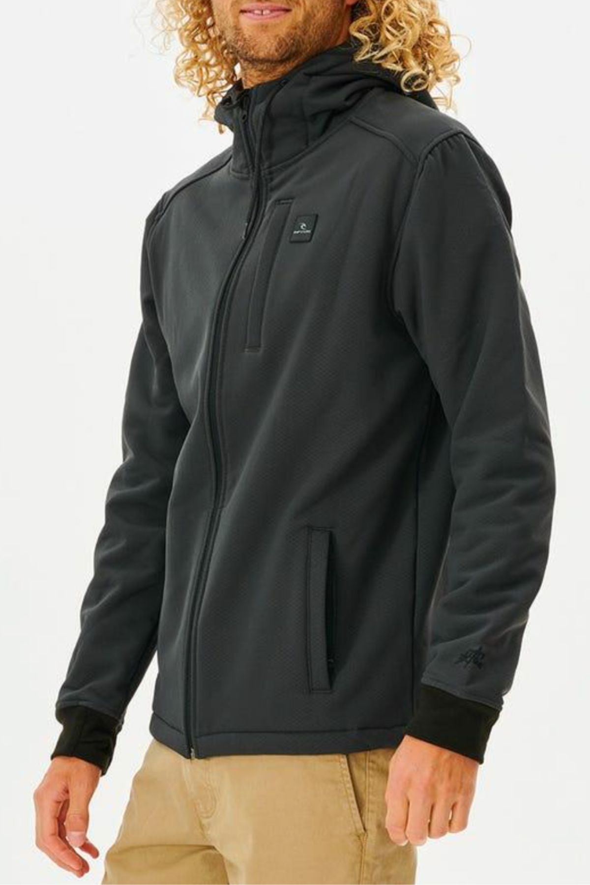 A person with long, curly blonde hair wears the black Rip Curl Anti Series Soft Tech Fleece jacket with a hood and beige pants. The water-resistant jacket has zippered pockets, a small chest logo, and is shown against a plain white background.