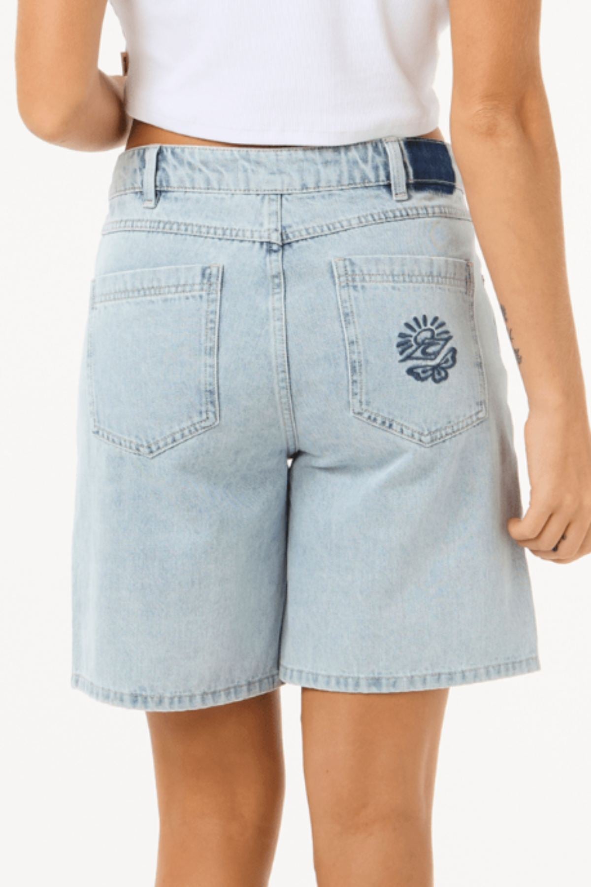 Model wearing Rip Curl Aggie Relaxed Denim Short in light blue with a white crop top, viewed from the back. The right back pocket features a small embroidered sun and wave design.