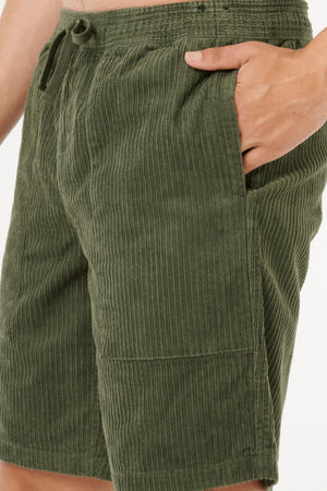 Someone wearing Rip Curl Classic Surf Cord Volley olive green cotton corduroy shorts, right hand in pocket, shown waist to mid-thigh against a white background, showcasing a vintage surf-inspired style.