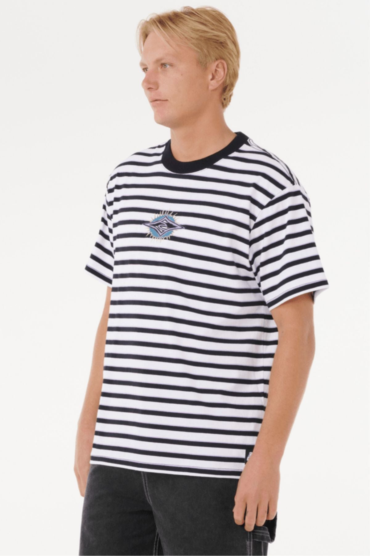 A person with blonde hair wears a black and white striped Rip Curl Raw Energy Stripey Tee with a small embroidered chest logo, standing against a plain white background.