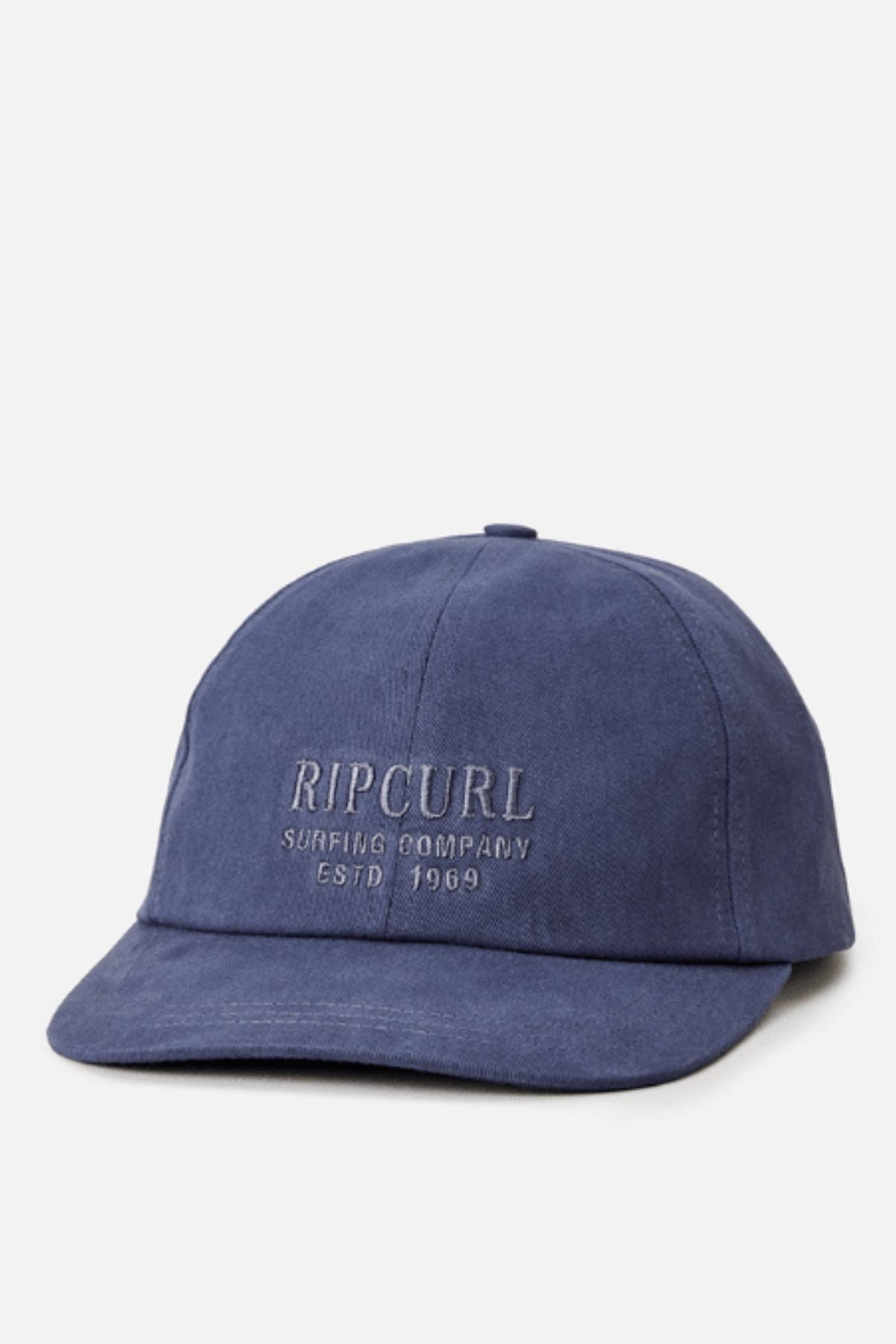 The Rip Curl Surf Staple Cap is blue with "RIP CURL SURFING COMPANY ESTD 1969" embroidered on the front. It features a six-panel design, curved brim, relaxed fit, and an adjustable strap for comfort.