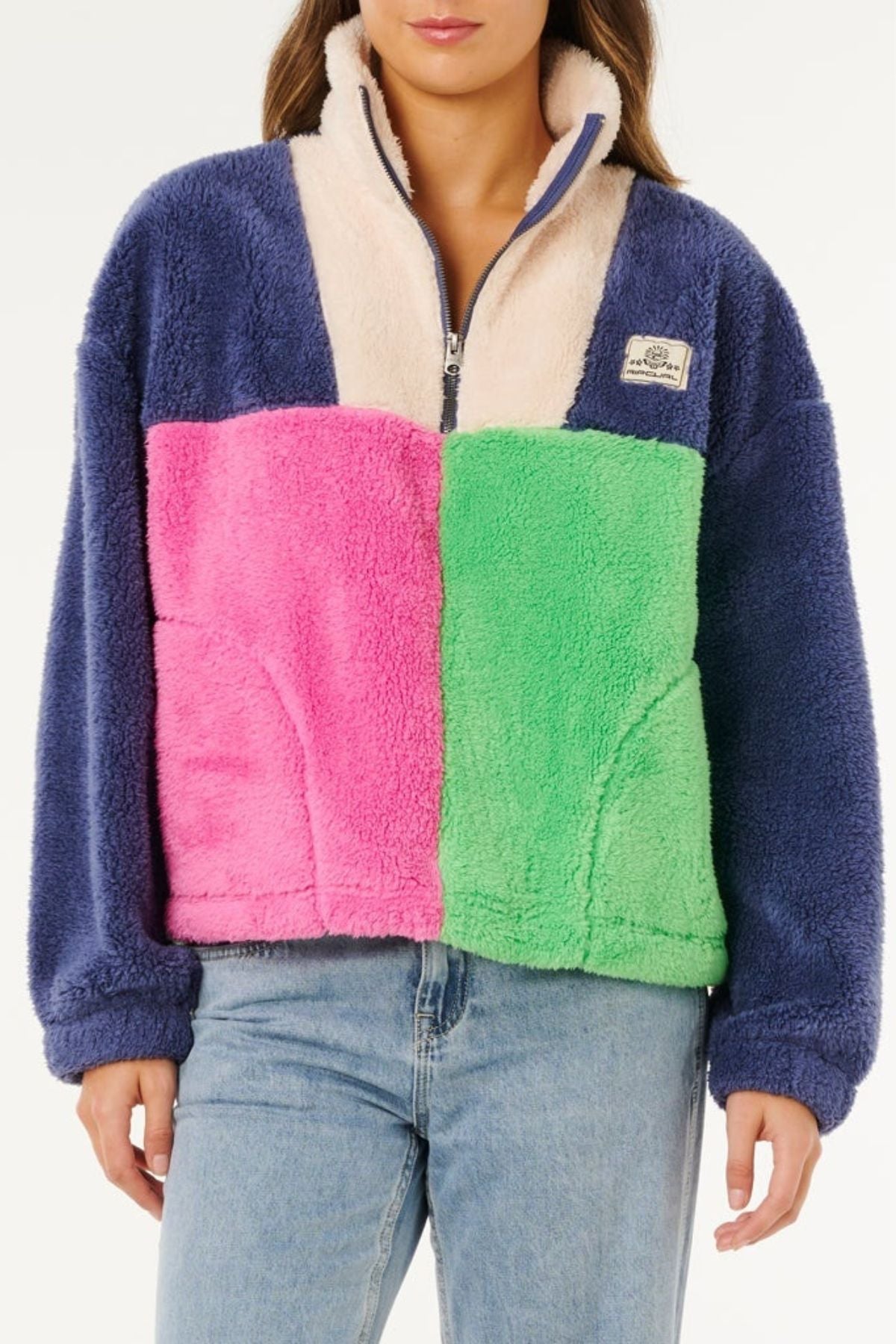 A woman wears a Rip Curl Block Party Polar Fleece with navy sleeves, cream top, pink left panel, green right panel, stylishly paired with blue jeans.