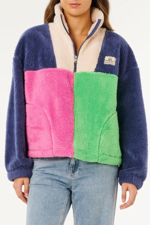 A woman wears a Rip Curl Block Party Polar Fleece with navy sleeves, cream top, pink left panel, green right panel, stylishly paired with blue jeans.