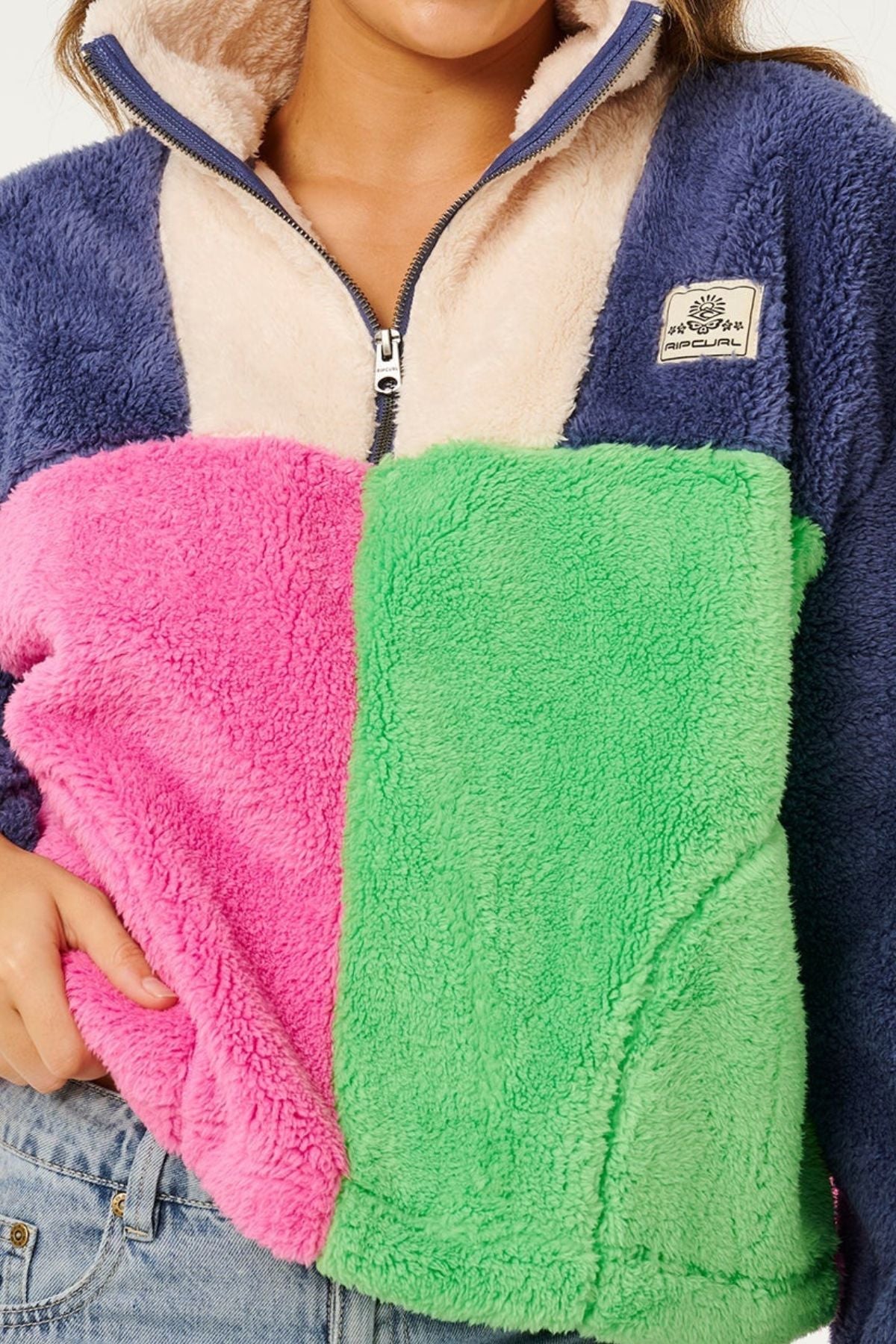 Wearing the Rip Curl Block Party Polar Fleece in navy, cream, pink, and green blocks, partially zipped with a chest patch and blue jeans—ideal for warmth in soft polyester microfleece.