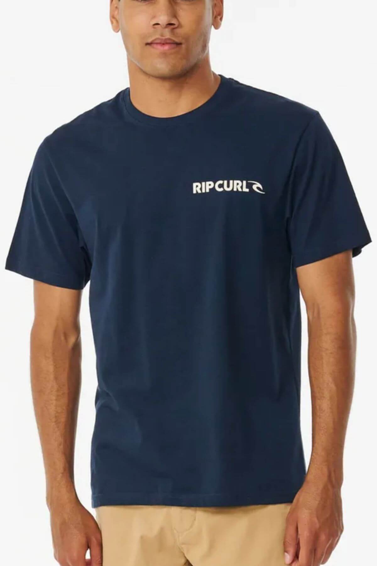 A person is wearing a navy blue Rip Curl Brand Icon Tee with beige pants. Made from 100% cotton, the tee showcases the Rip Curl logo on the chest, styled in a standard fit and contrasted against a plain white background.