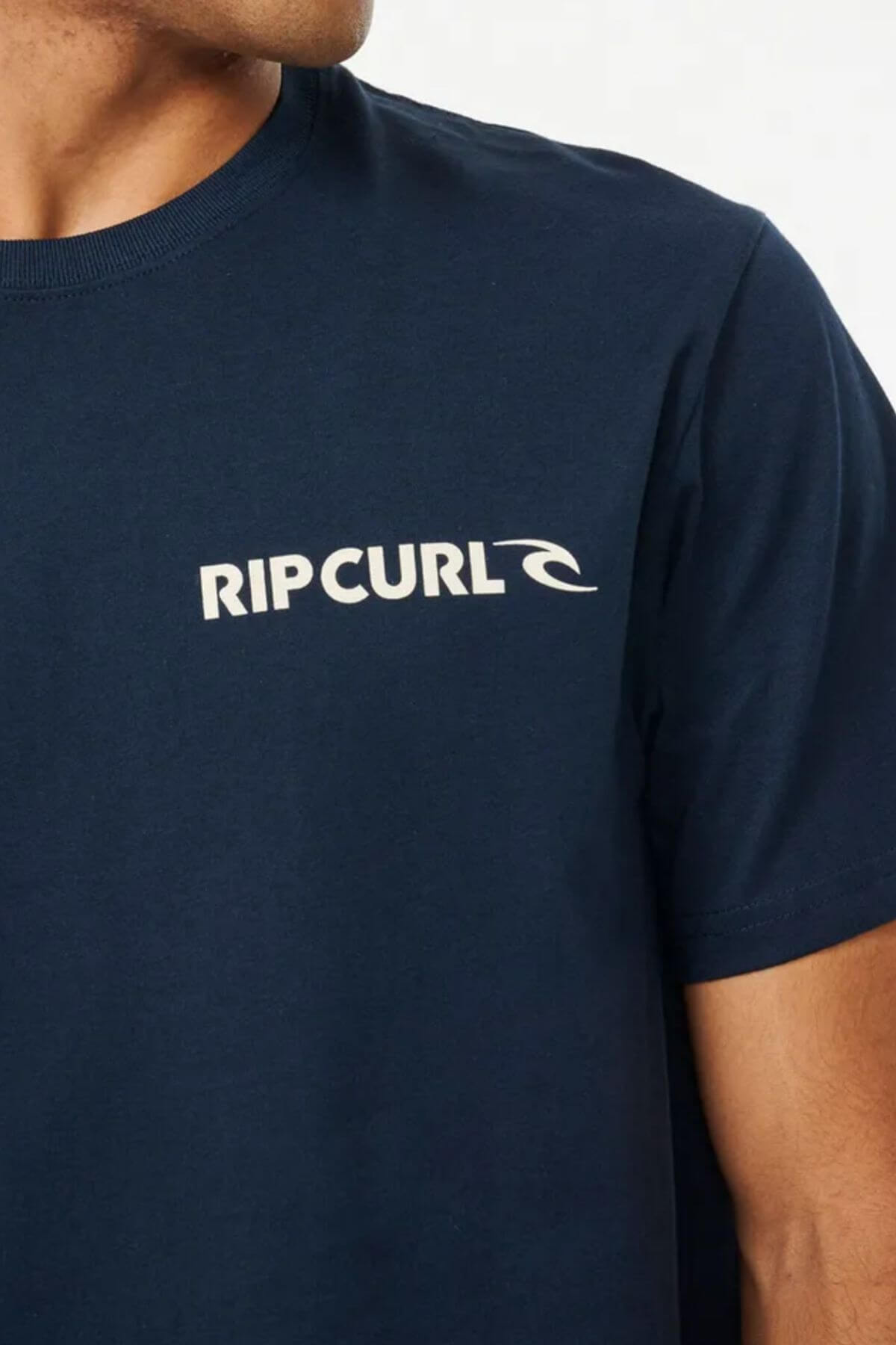 A close-up shows a person wearing the Rip Curl Brand Icon Tee in navy blue. The tee features the "RIP CURL" logo in white on the left chest. This standard-fit shirt, crafted from 100% cotton, is showcased as the person's face remains partially out of frame.