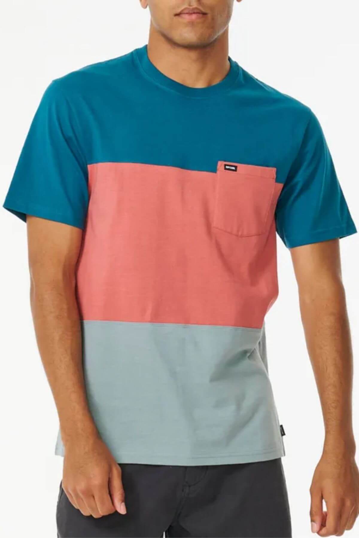 A man wears the Rip Curl Divided Tee, a 100% cotton crew neck t-shirt with teal, coral pink, and light gray color blocks plus a chest pocket, paired with dark shorts.
