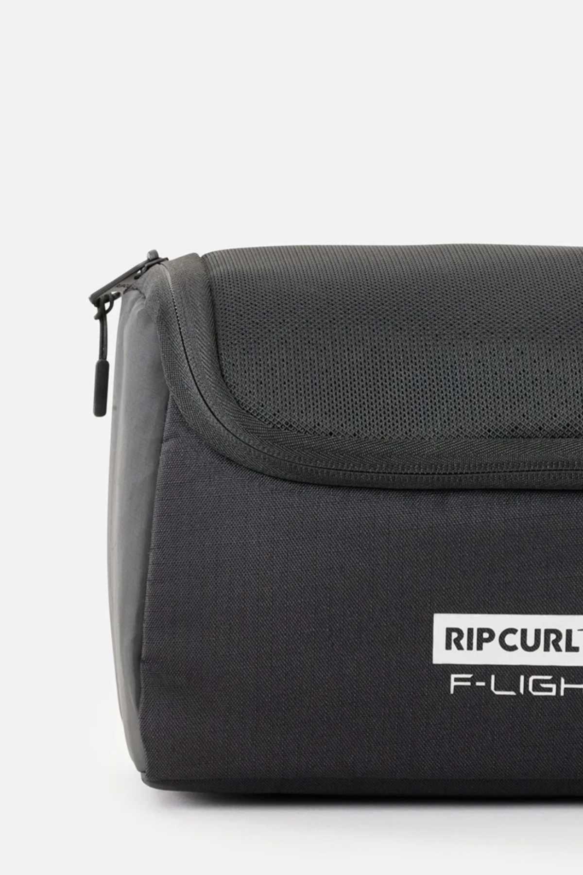 A close-up of a black Rip Curl F-Light Toiletry bag on a white background showcases its multiple compartments, zipper closure, ventilated mesh section, and prominently printed side logo.