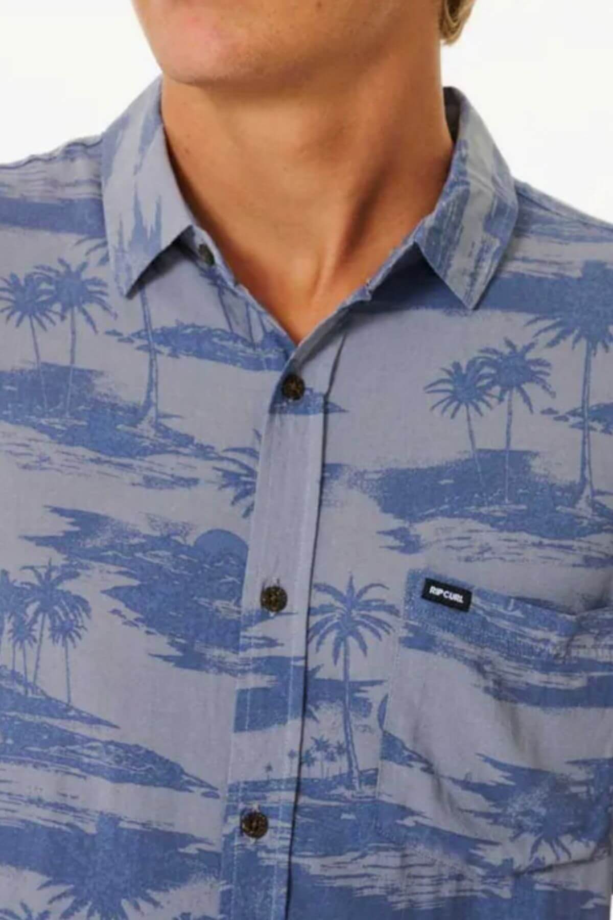 A person wears a Rip Curl Party Pack Short Sleeve Shirt in blue, featuring a tropical palm tree and island pattern, collar, chest pocket with black and white label; their face is partially hidden.