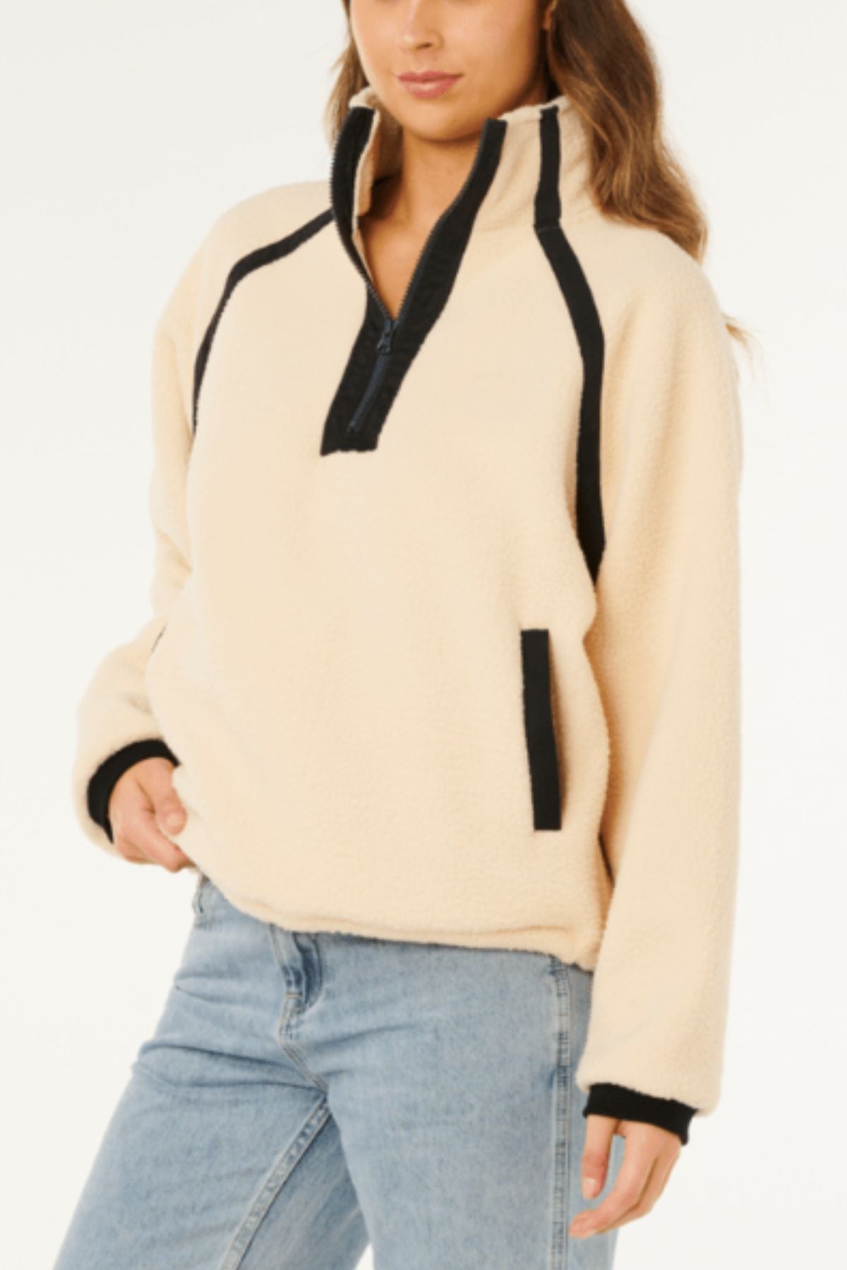 A woman wears the Rip Curl Surf Club Heritage Polar, a cream-colored polar fleece pullover with black trim and zipper, high collar, and long sleeves, paired with light blue jeans against a plain white background.