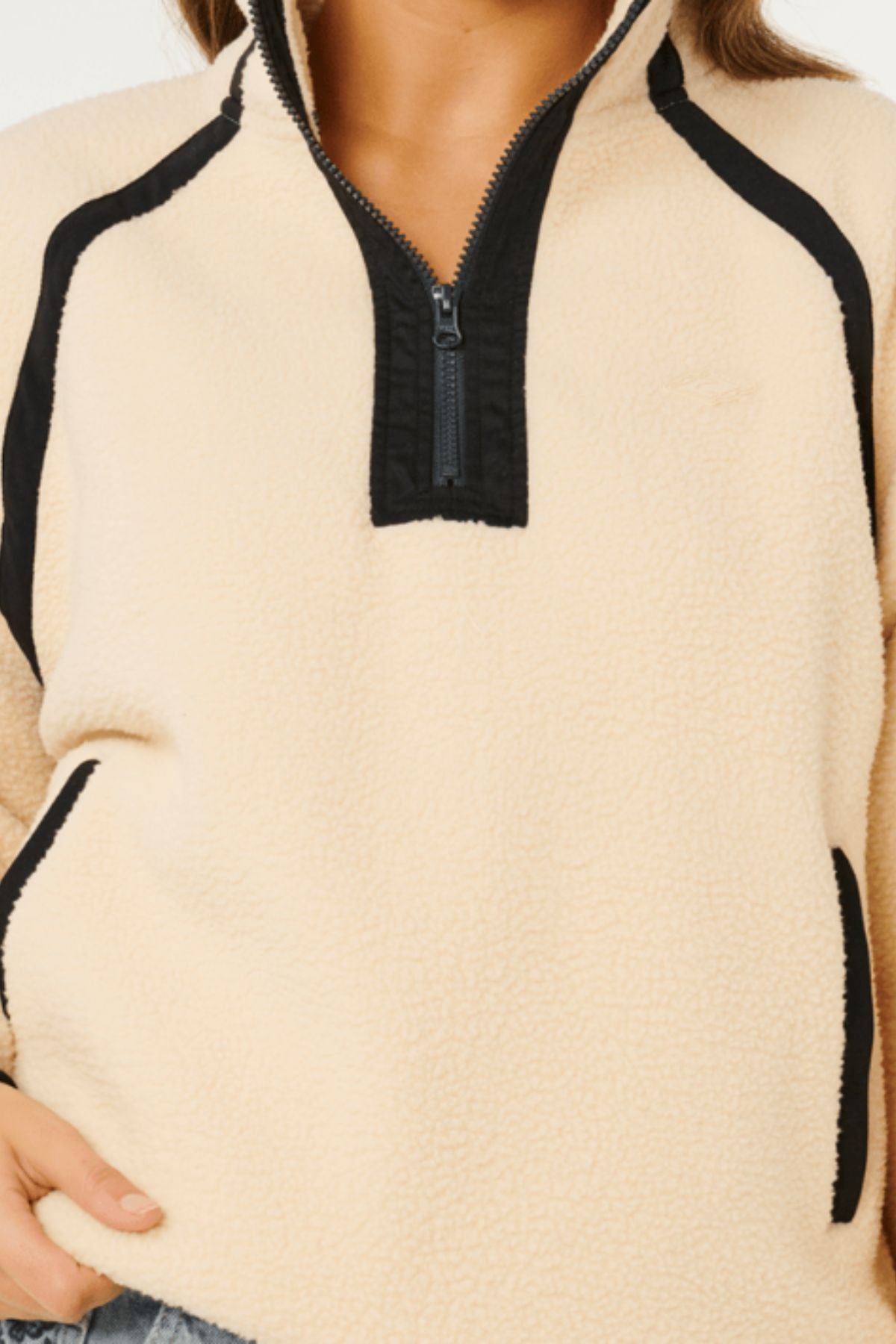 A person wearing the Rip Curl Surf Club Heritage Polar—a cream-colored fleece pullover with black trim, a zip collar, and black-trimmed pockets. Only their lower face and upper body are shown.