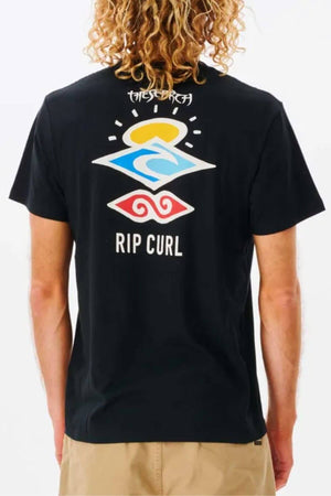 A curly-haired individual wears a black Rip Curl Search Icon Tee showcasing vibrant back graphics with "RIP CURL" text, sun, waves, and abstract shapes, perfectly paired with beige pants.