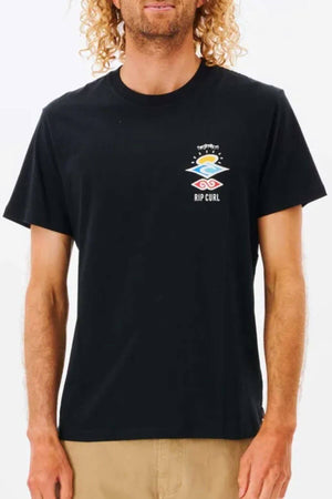 Wearing a black Rip Curl Search Icon Tee made from soft cotton, the person displays a vibrant graphic logo featuring geometric shapes and "Rip Curl" text. Paired with beige pants and long curly hair, they capture a relaxed vibe.