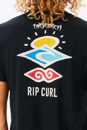 A person with curly hair is seen from behind wearing a Rip Curl Search Icon Tee, a black cotton T-shirt featuring colorful abstract shapes and "RIP CURL" in white.
