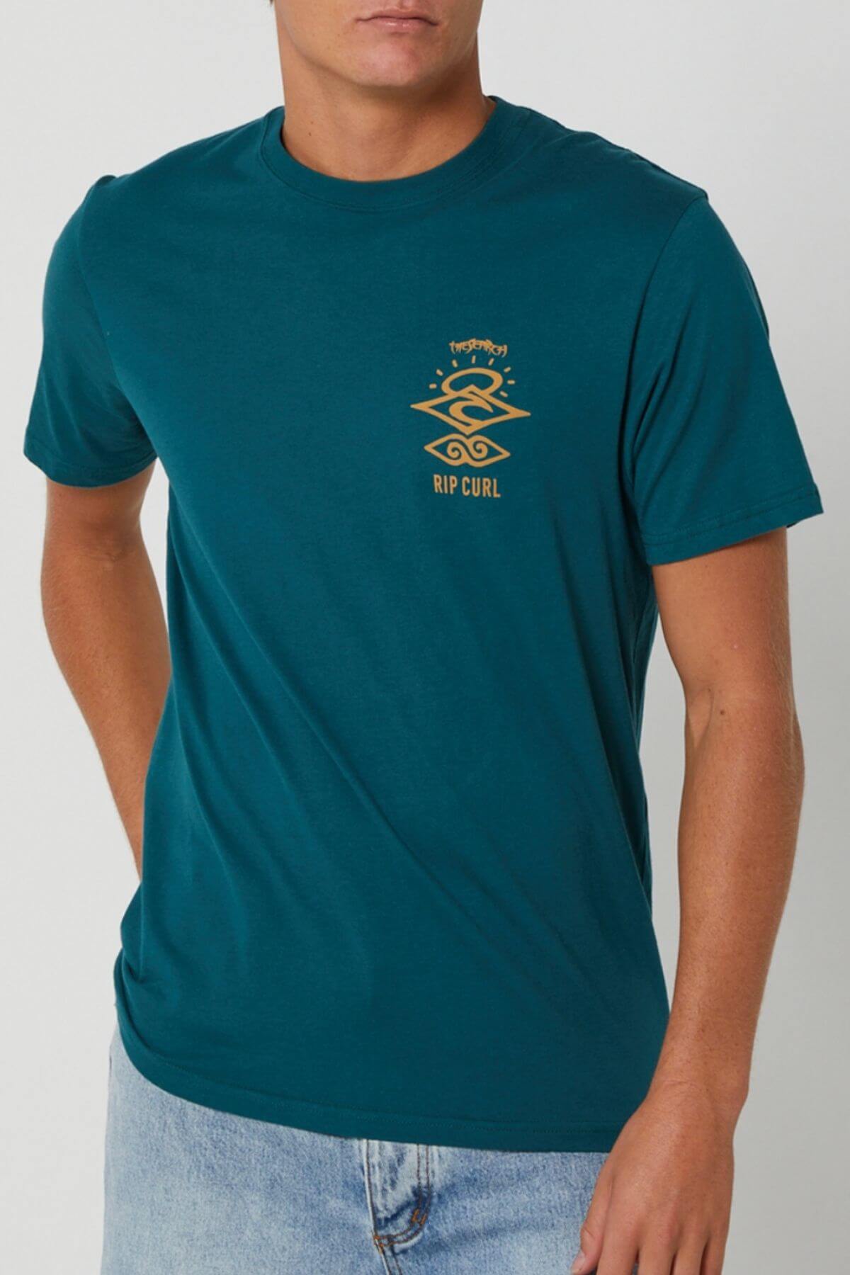 Someone wearing the teal Rip Curl Search Icon Tee, which has a small yellow logo and design on the left chest, pairs it with light blue jeans while standing against a plain light background.