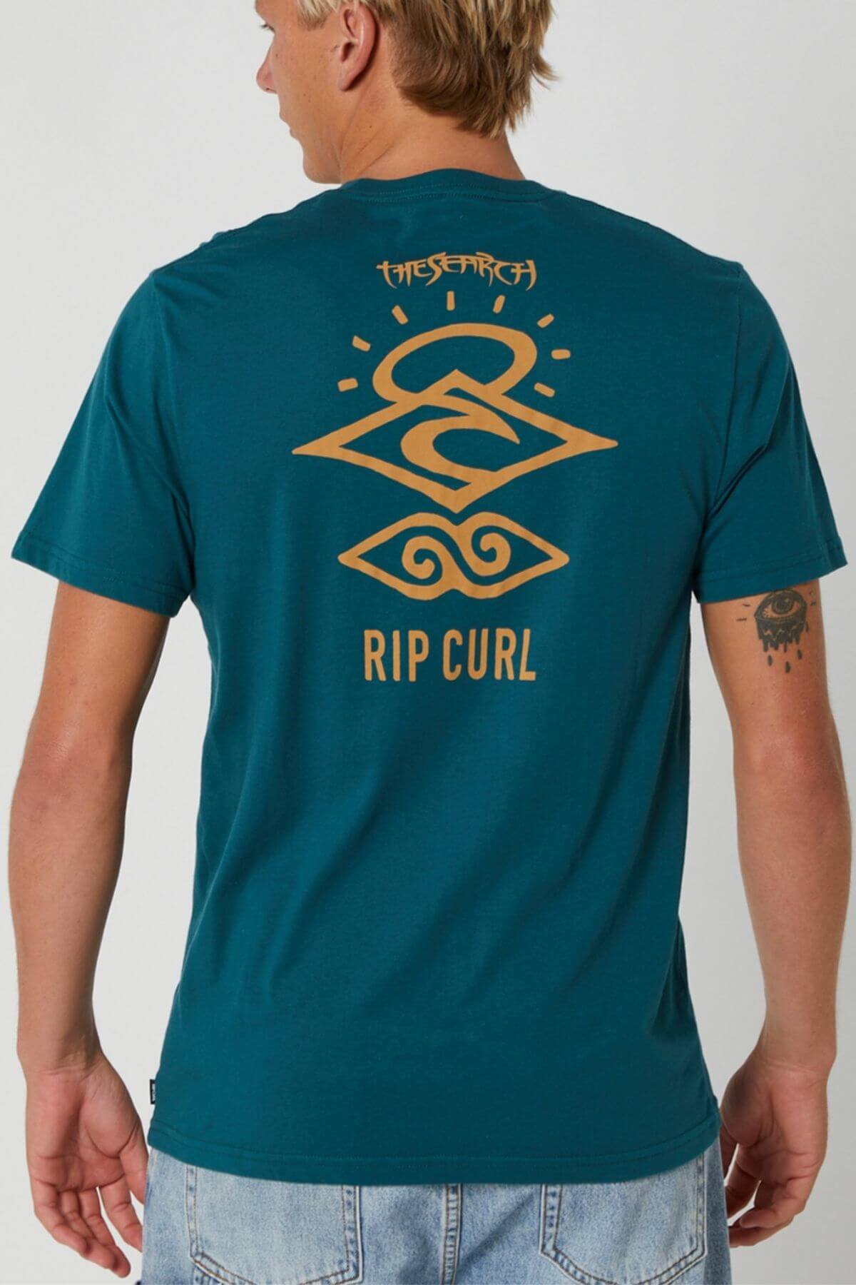 Someone faces away from the camera wearing the Rip Curl Search Icon Tee—a teal cotton shirt with a bold tan geometric graphic and "THE SEARCH" plus "RIP CURL" printed on the back.