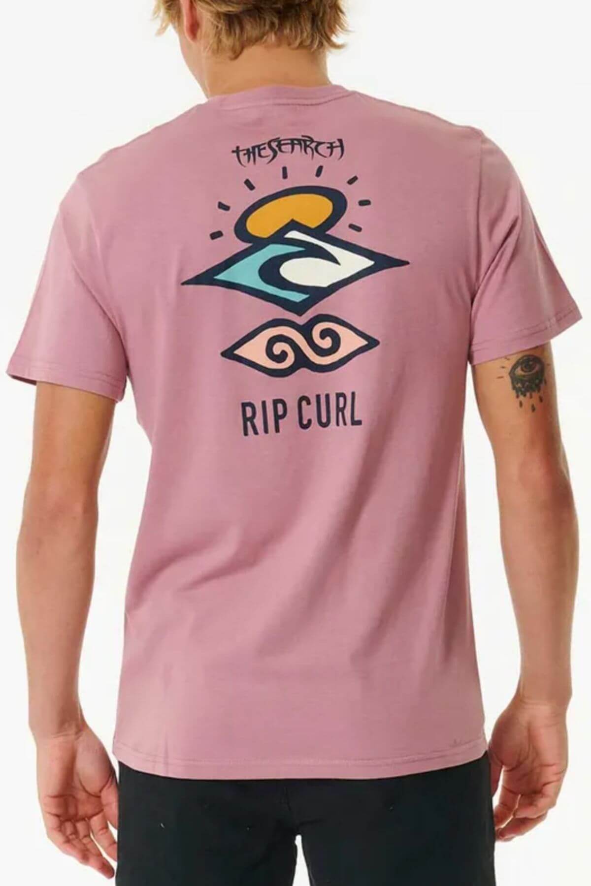 Wearing a pink Rip Curl Search Icon Tee featuring abstract shapes, a wave, and the "THE SEARCH" text on the back, a person with short hair exudes a laid-back vibe, captured turned away in this comfy cotton tee.