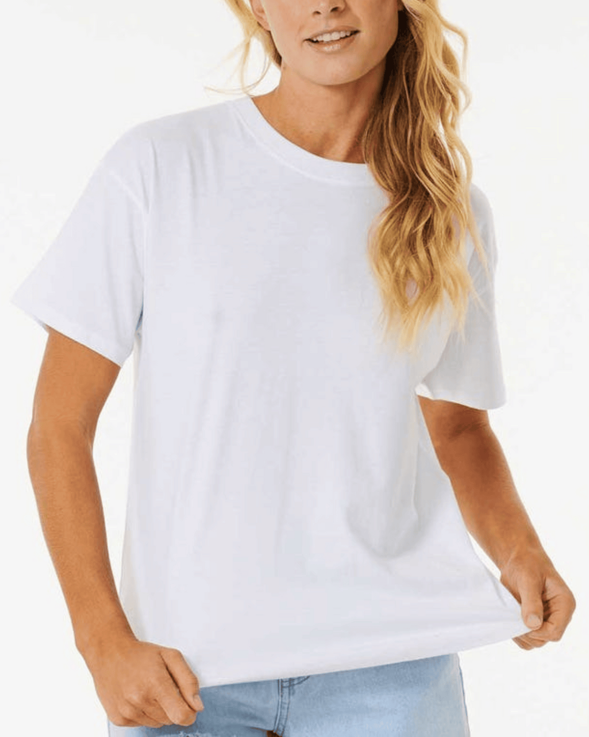 A person with long, wavy hair is wearing a Rip Curl Classic Relaxed Tee in Optical White paired with light blue jeans, exuding a casual and laid-back vibe. They are standing against a white background, gently holding the side of their cotton jersey T-shirt.