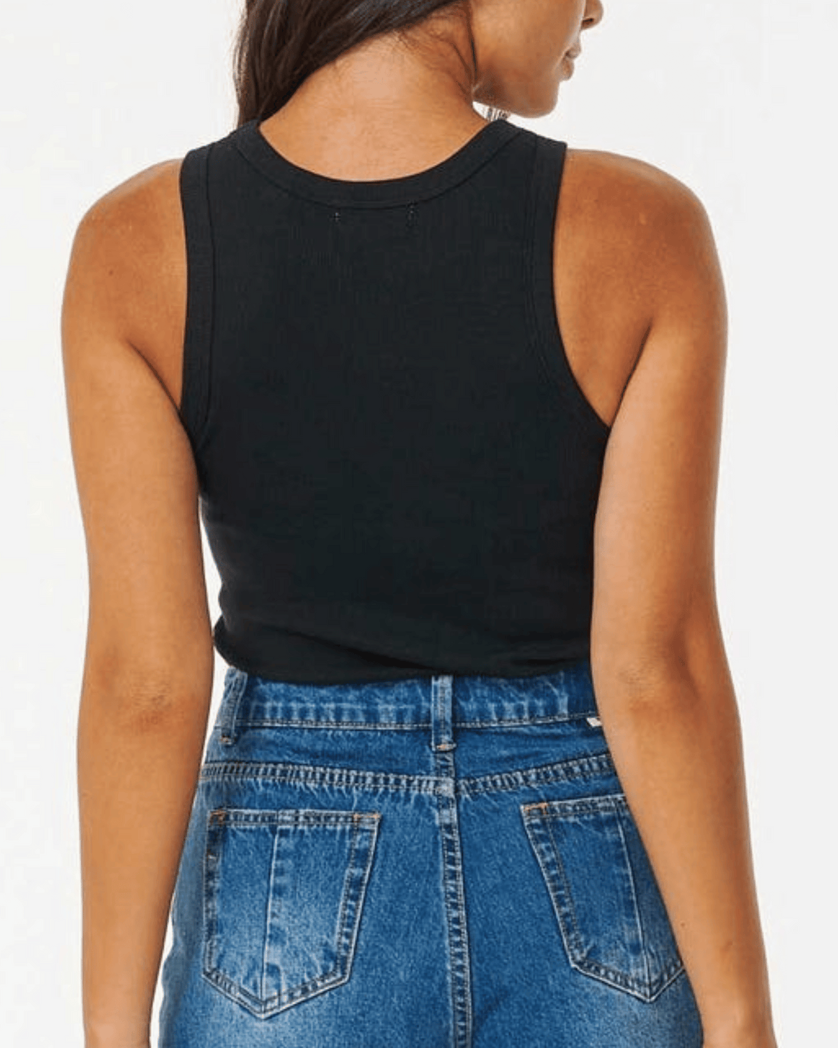 A woman with long brown hair is shown from the back, wearing a black Rip Curl Classic Ribbed Tank and high-waisted blue jeans.